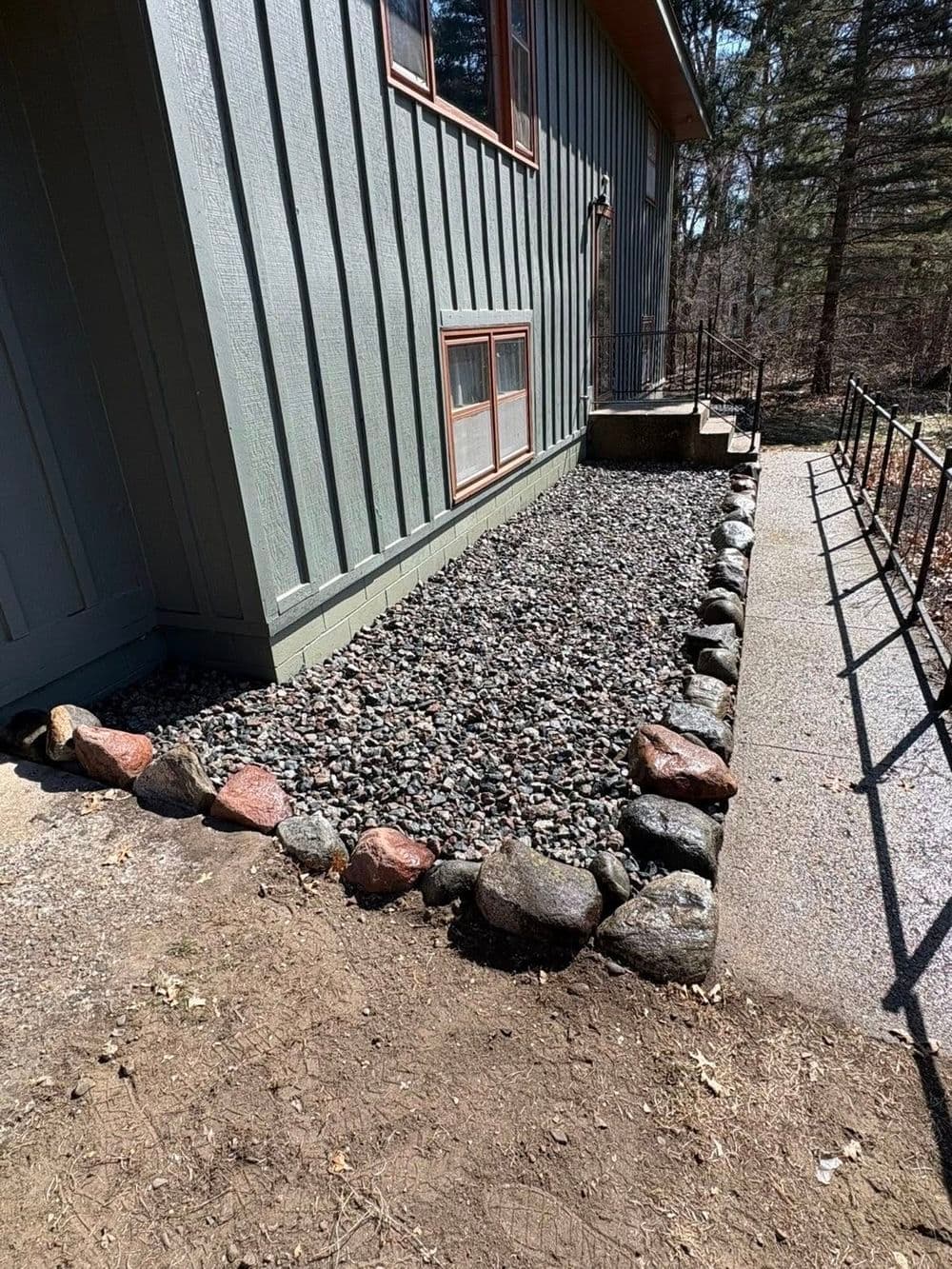 Gravel landscaping with rock border beside a green house in a wooded area.