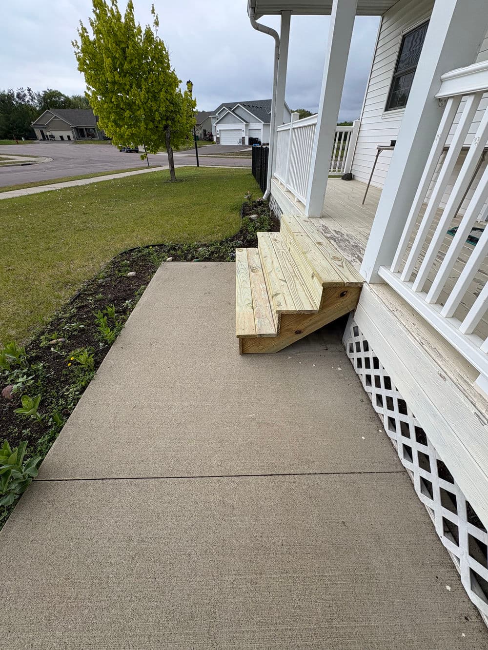 Wooden steps leading to a porch with concrete sidewalk and landscaped yard.