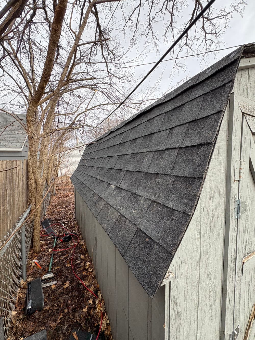 Gray shed with asphalt shingles beside bare trees and garden tools in a backyard setting.