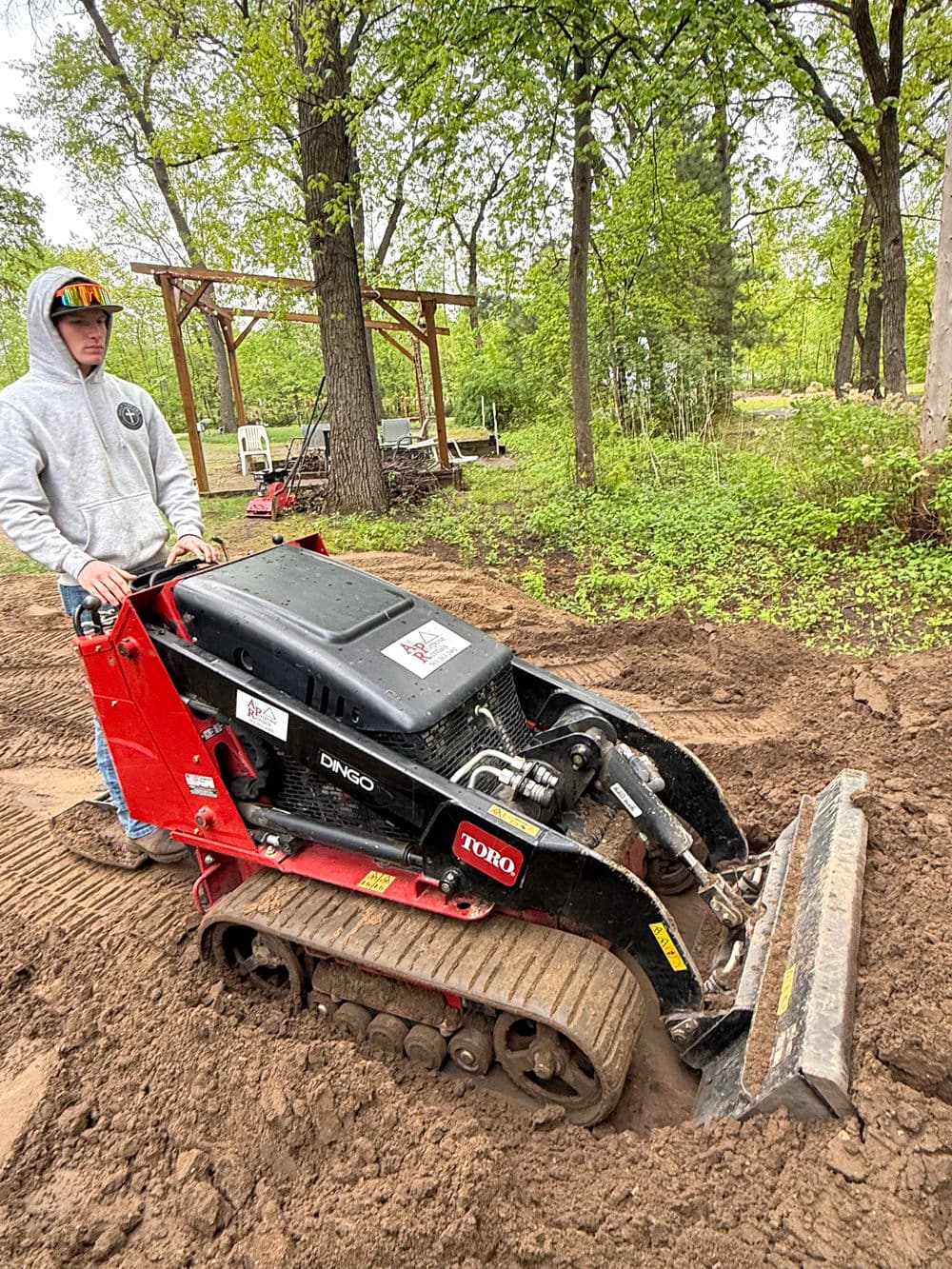 Person operating a Toro Dingo compact equipment in a wooded area, preparing the ground.