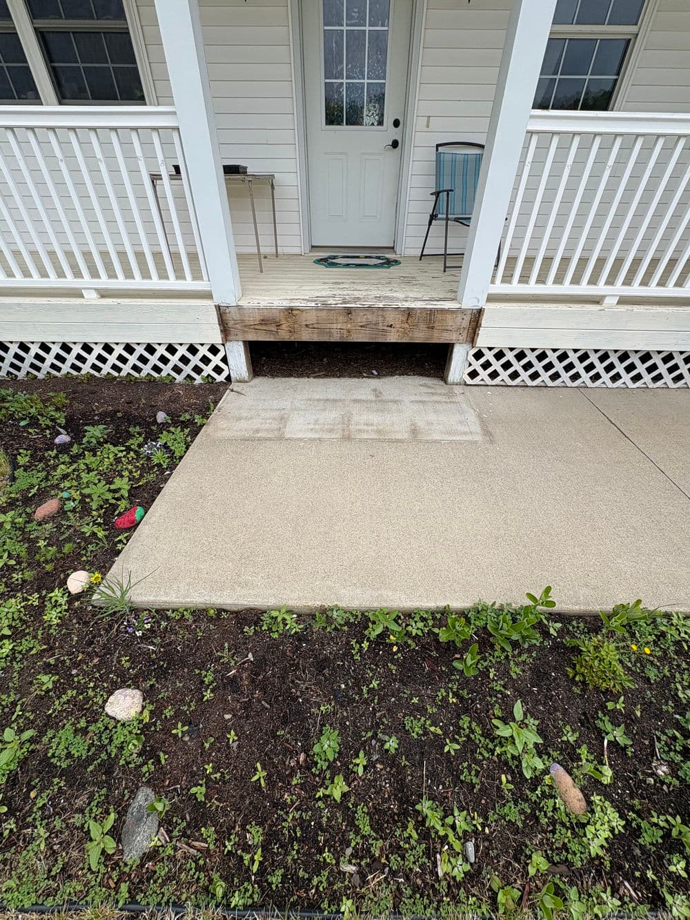 Front porch entrance with a wooden step and concrete walkway, surrounded by greenery.