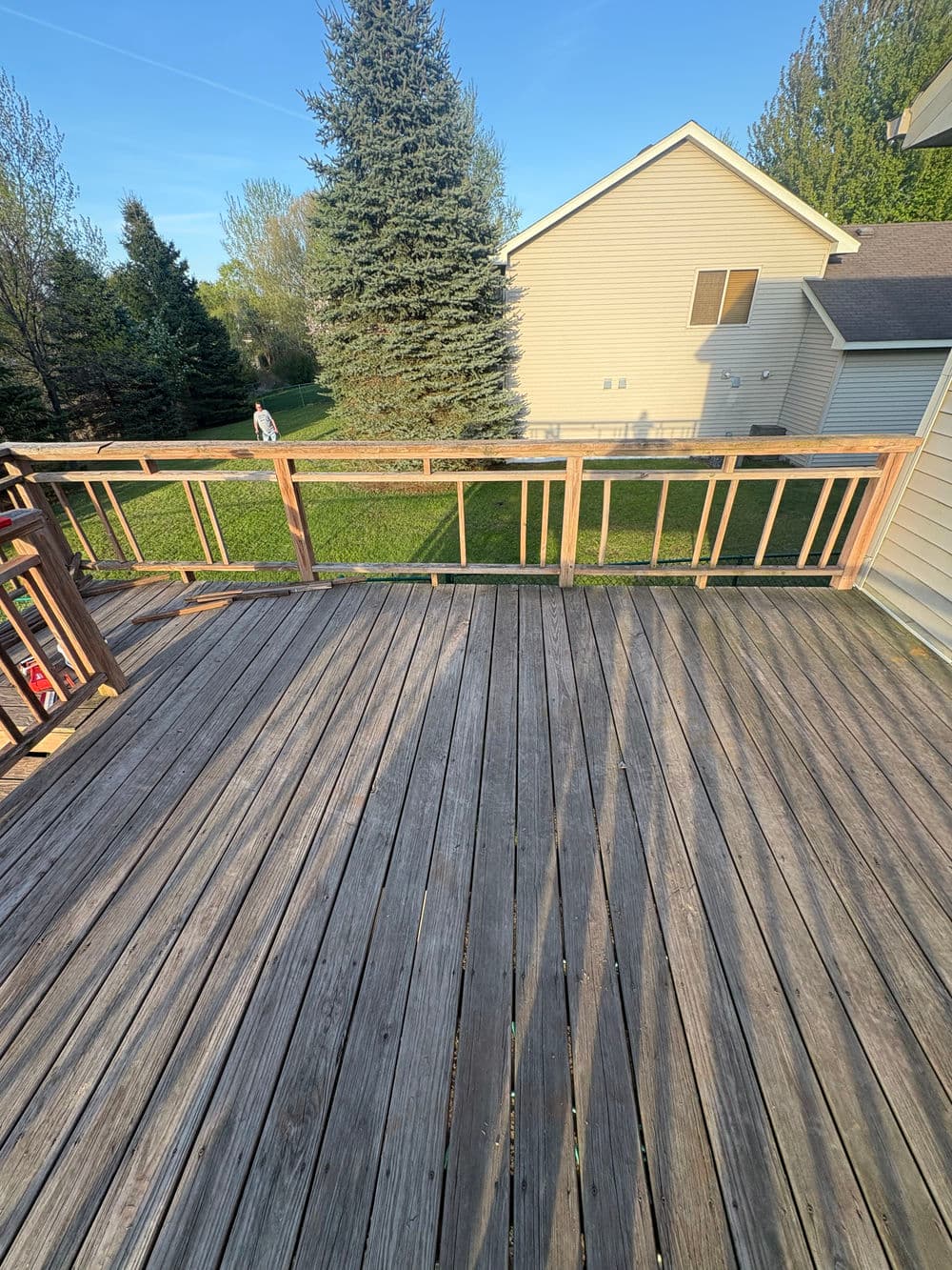 Deck view with wooden railing, grass lawn, trees, and neighboring house under clear blue sky.