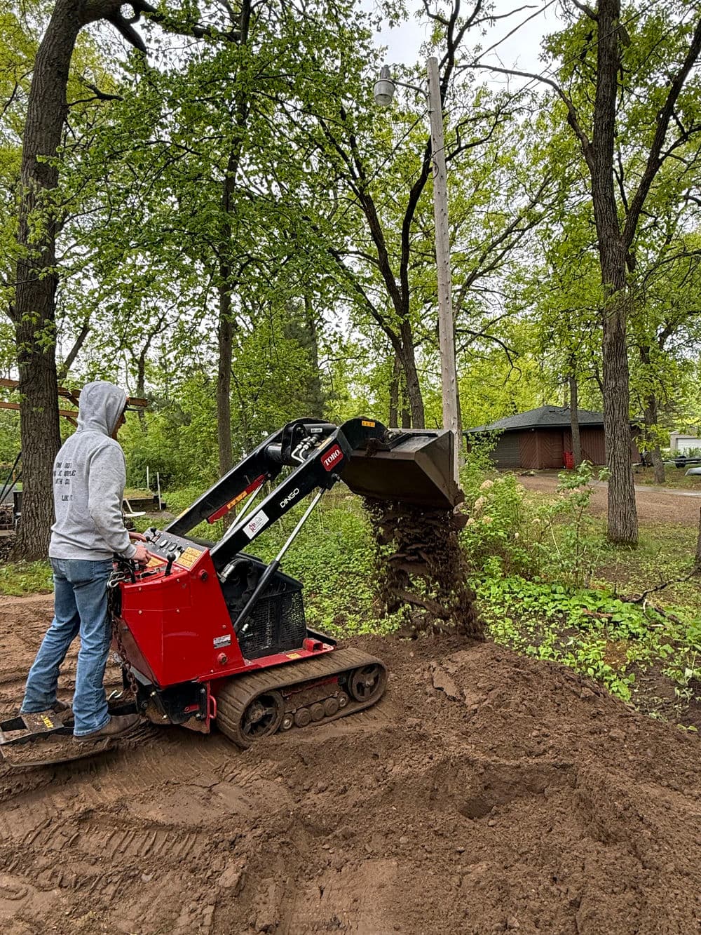 Person operating a mini skid steer loader to move soil in a wooded area.